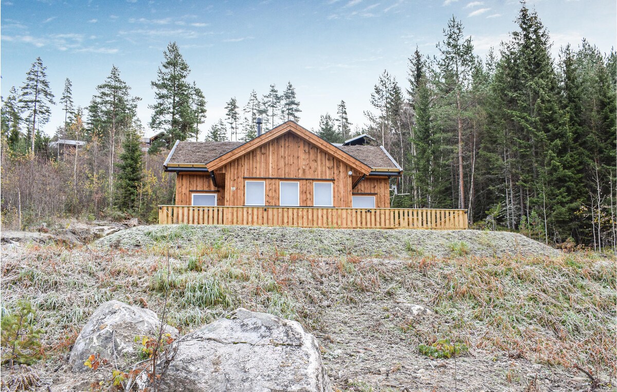 A wooden vacation home is set against a backdrop of tall trees and open landscape. Large windows provide views of the surroundings. A wooden railing encloses the front area, which is covered in low grass and scattered rocks.