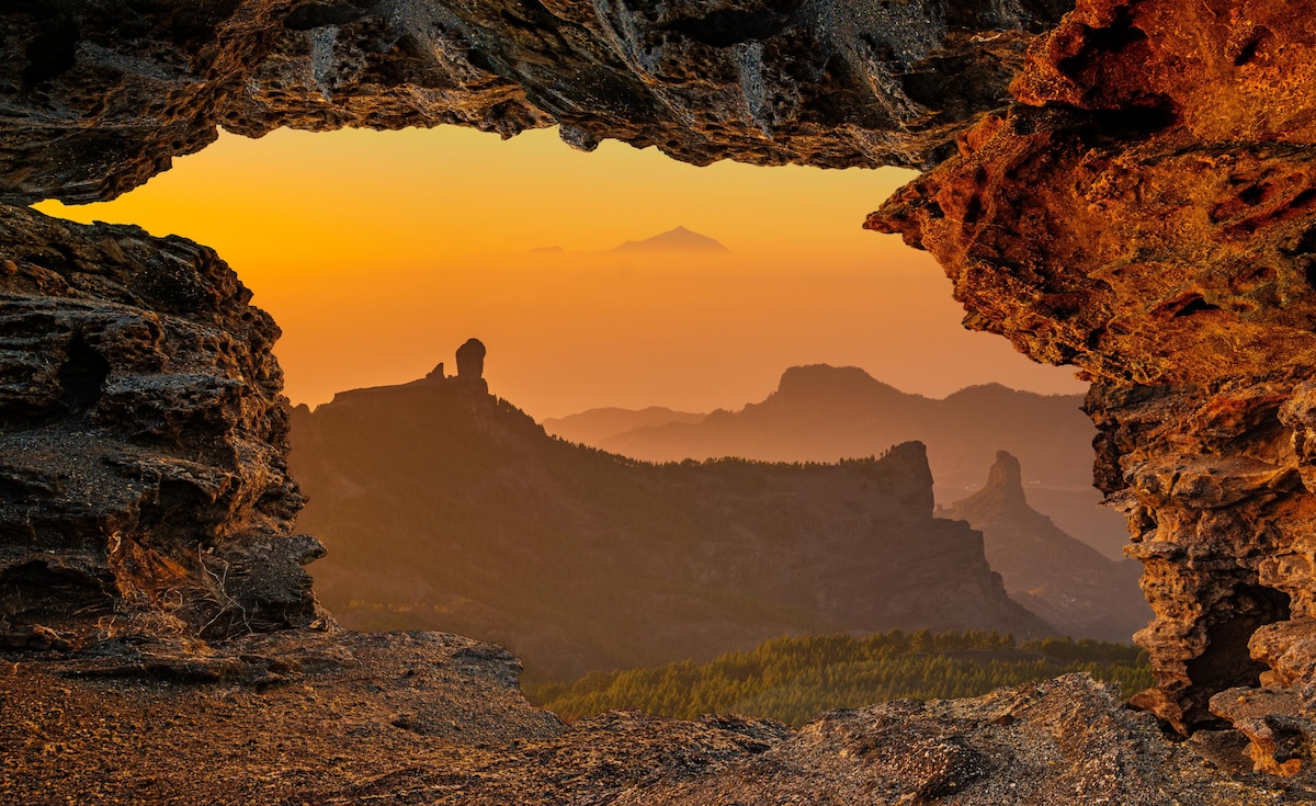 A natural rock formation frames a stunning view of layered mountains at sunset. Warm orange and gold hues illuminate the landscape, highlighting the silhouette of distant peaks against the horizon, creating a serene and picturesque scene.
