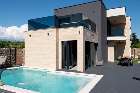 The modern villa exterior showcases a well-designed layout with a private pool and terrace area. Glass railings elevate the balcony space, while potted plants add a touch of greenery. The surrounding environment features a blend of natural elements, complementing the villa's aesthetic.