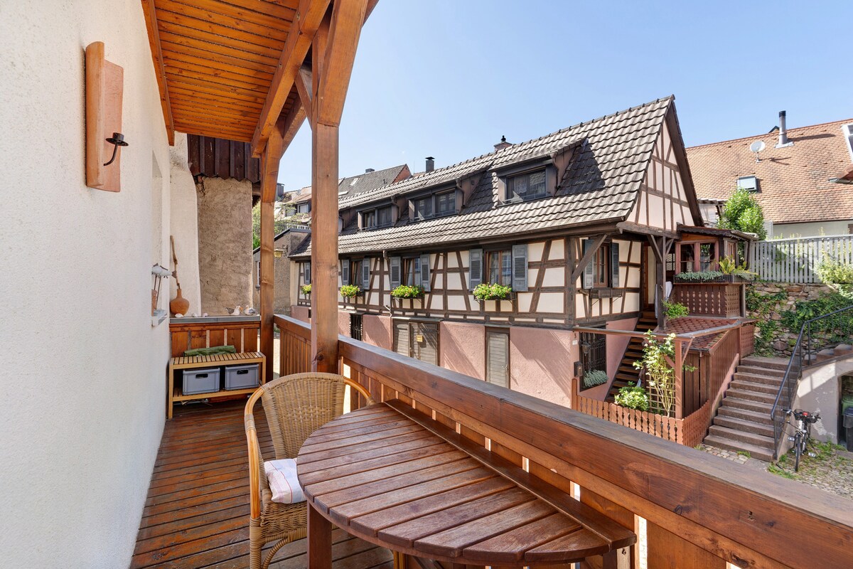 A private balcony features wooden flooring and a round table with one chair, overlooking traditional half-timbered houses. The space is bathed in natural light, creating an inviting area for relaxation. Lush greenery is visible between the buildings, enhancing the tranquility of the scene.