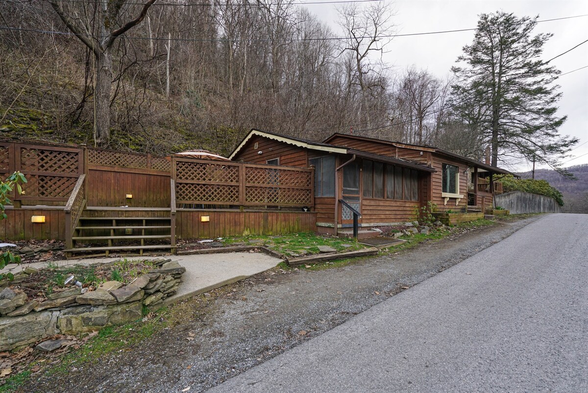 A rustic cabin exterior is set against a backdrop of trees, featuring wooden siding and a spacious front porch. The structure is surrounded by pathways and landscaping, adding to the natural setting. Street access is visible in the foreground, providing a clear view of the cabin's inviting entrance.