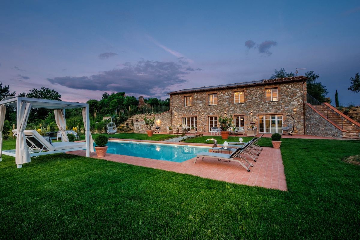 The outdoor area features a private swimming pool surrounded by lush green grass. A stone farmhouse with multiple windows is visible in the background, complemented by neatly arranged plants and lounge chairs. Soft lighting enhances the serene atmosphere as the sky transitions into twilight.