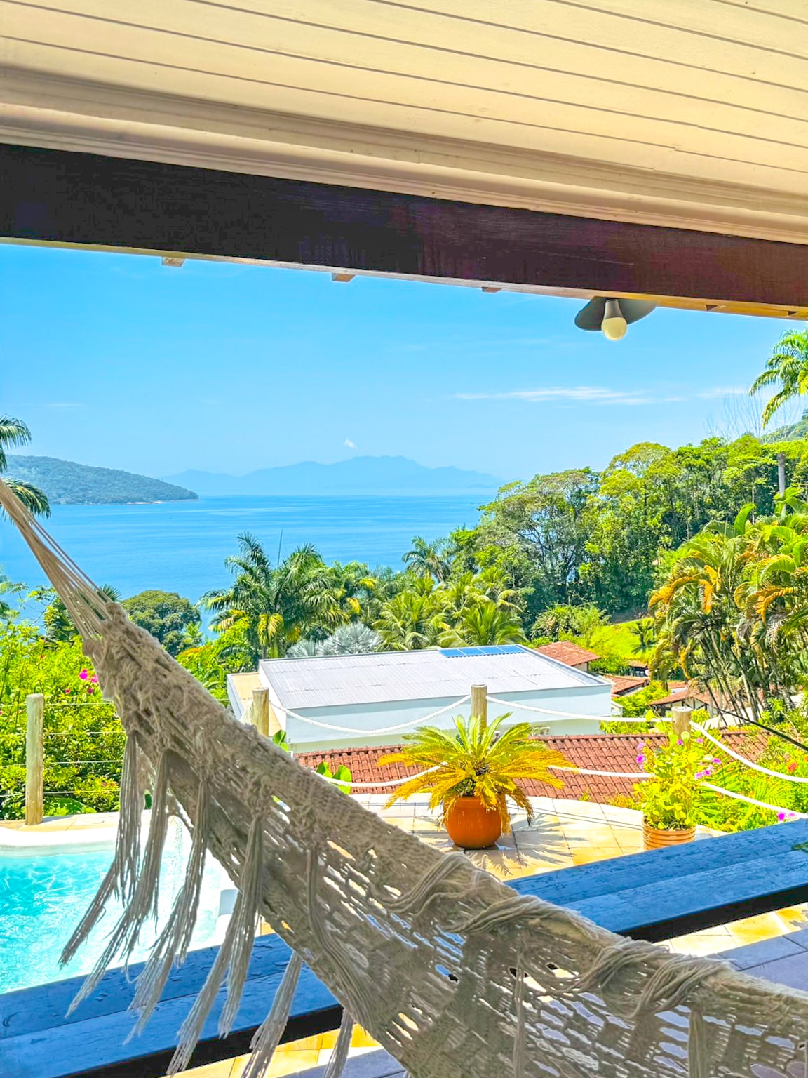 A view of the serene sea and lush greenery is captured from a balcony. A hammock hangs invitingly, with vibrant tropical plants visible in the foreground, and the shimmering water blending with distant hills under a bright sky.