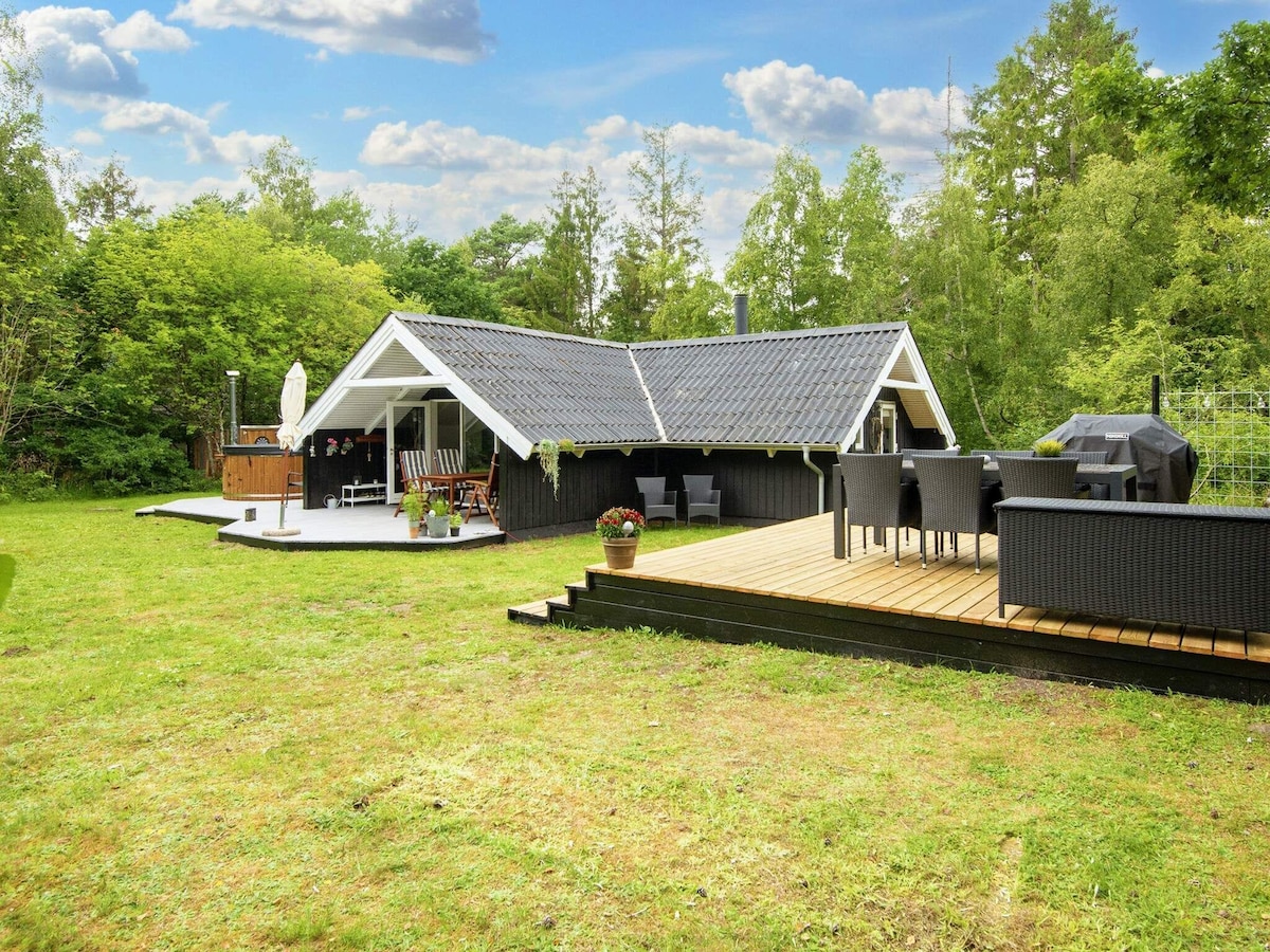 A spacious holiday home is set in a green landscape, featuring a large deck area with outdoor seating and a grill. The house has a sloped roof with a modern design, surrounded by trees and a manicured lawn, creating a serene outdoor setting.