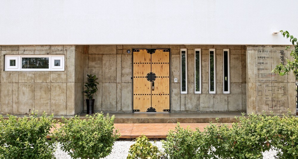 The entrance features a large wooden door with intricate detailing, flanked by narrow vertical windows. A small plant stands in a pot beside the door, and a pathway made of wooden planks leads toward the well-maintained garden area with low hedges.