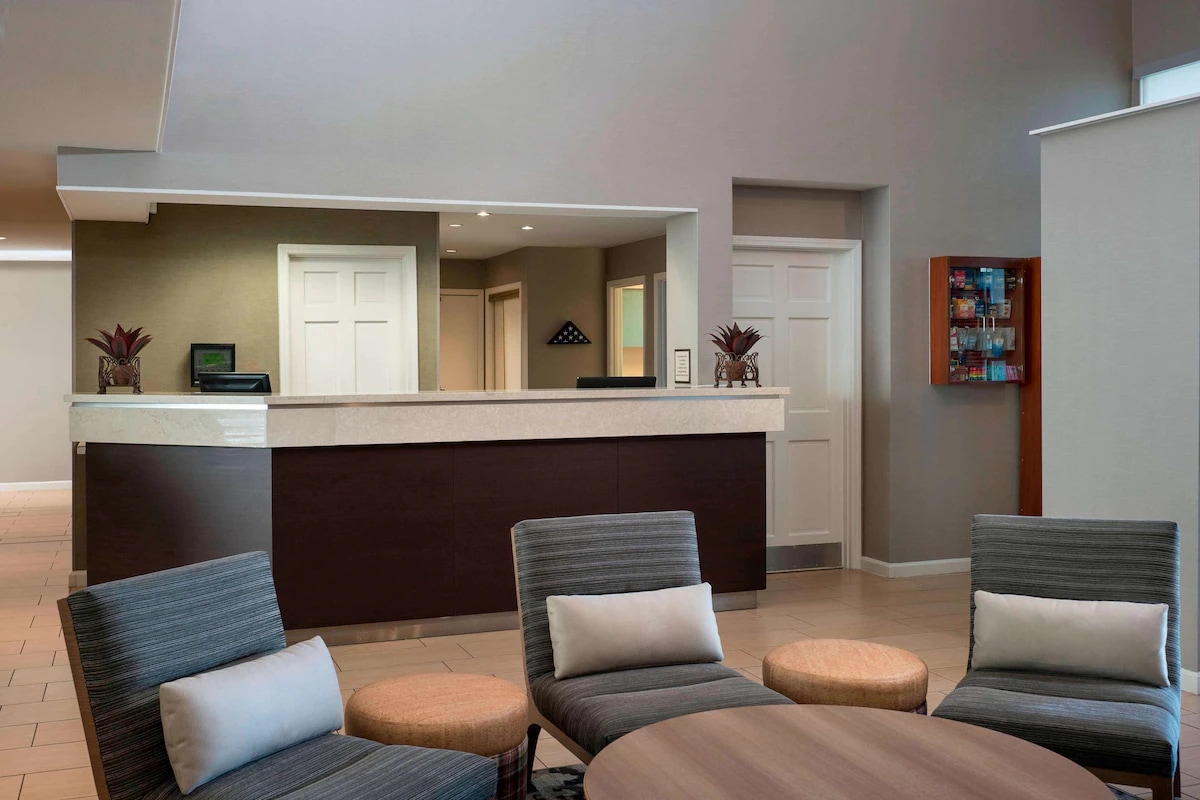The reception area features a modern front desk made of light-colored stone, complemented by stylish seating arrangements. Soft-colored chairs and round tables create a welcoming space for guests. Natural light fills the area through nearby doors, enhancing the openness.