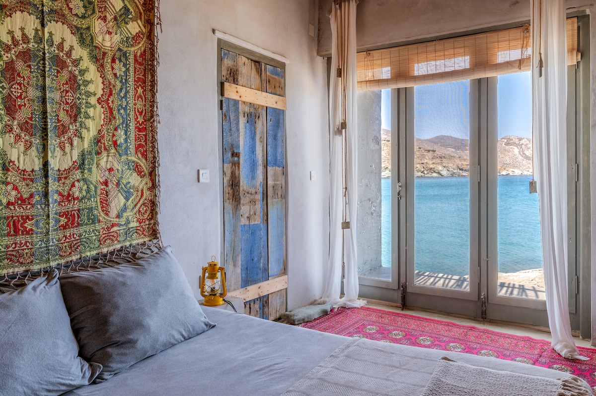A serene bedroom is featured with a colorful wall tapestry behind a spacious bed. Large glass doors open to a stunning view of the Aegean Sea, flooding the room with natural light. A lantern sits on a small bedside table, enhancing the tranquil ambiance.