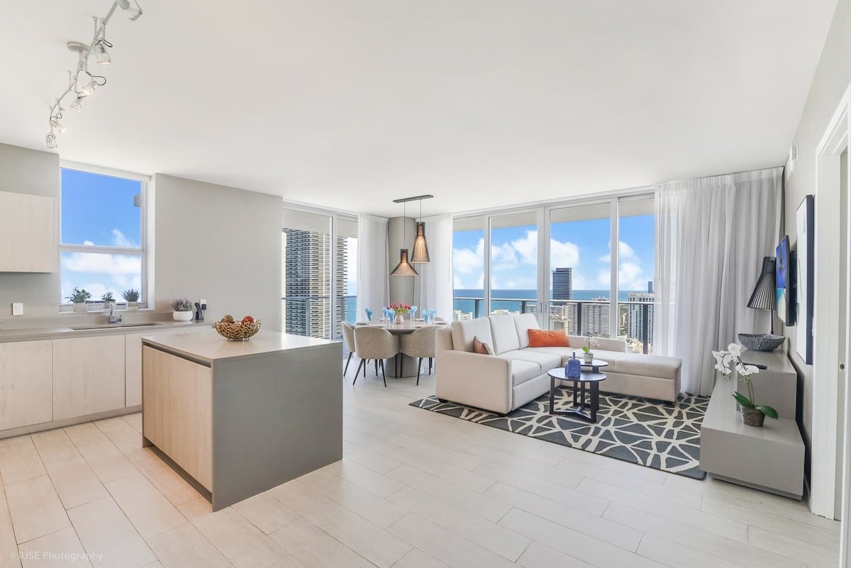 An open living area is brightened by natural light from large floor-to-ceiling windows, showcasing ocean views. A comfortable sectional sofa is placed on a patterned rug, complemented by a dining table set for six. The kitchen features modern cabinetry and an island for additional workspace.