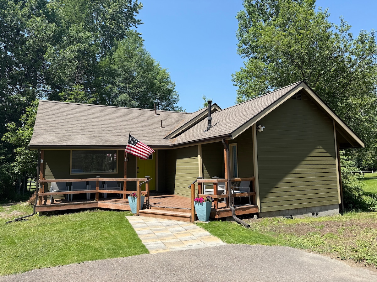 The exterior of a green house is displayed, featuring a covered porch with a wooden railing. An American flag is mounted, and flower pots are positioned on the steps. The surrounding lawn is well-maintained, and trees provide a natural backdrop.