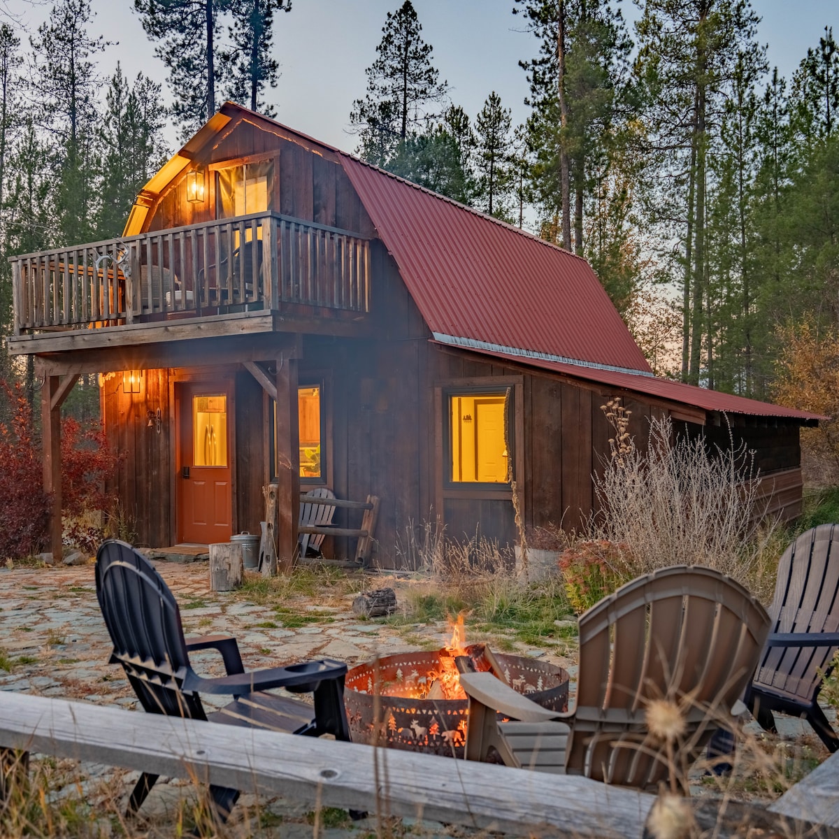 The exterior of a rustic cabin is framed by tall trees and a stone patio. Soft light glows from the windows and a balcony above. Four comfortable chairs are arranged around a fire pit, inviting relaxation and warmth during evenings outdoors.