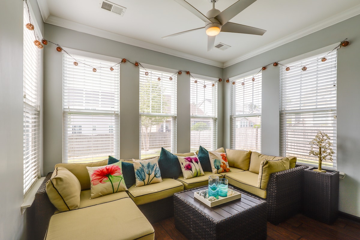 A sunlit sunroom is designed with a large sectional couch adorned with colorful pillows. Large windows, framed in white, provide ample natural light while sheer curtains softly diffuse it. A glass-top coffee table sits at the center, featuring decorative elements.