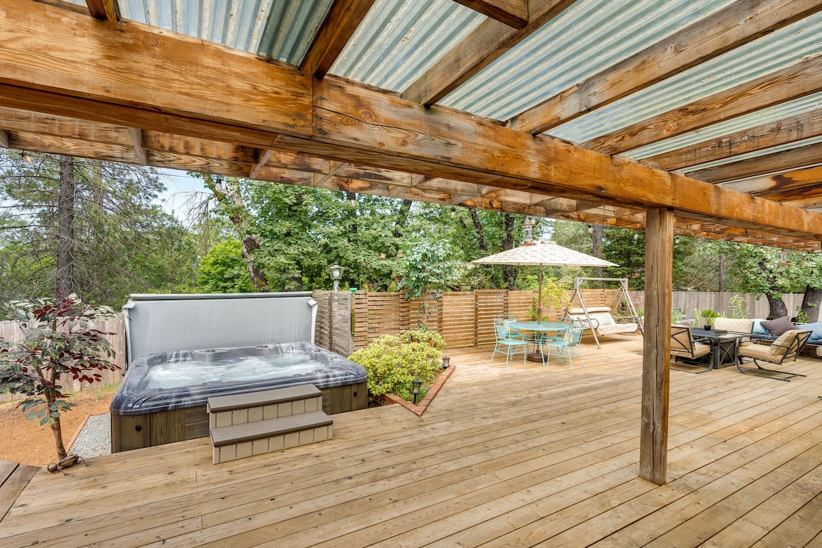 An outdoor living space features a private hot tub beside a spacious wooden deck. A shaded dining area is visible, adorned with an umbrella and surrounded by seating. Lush greenery provides privacy around the yard, contributing to a serene atmosphere.