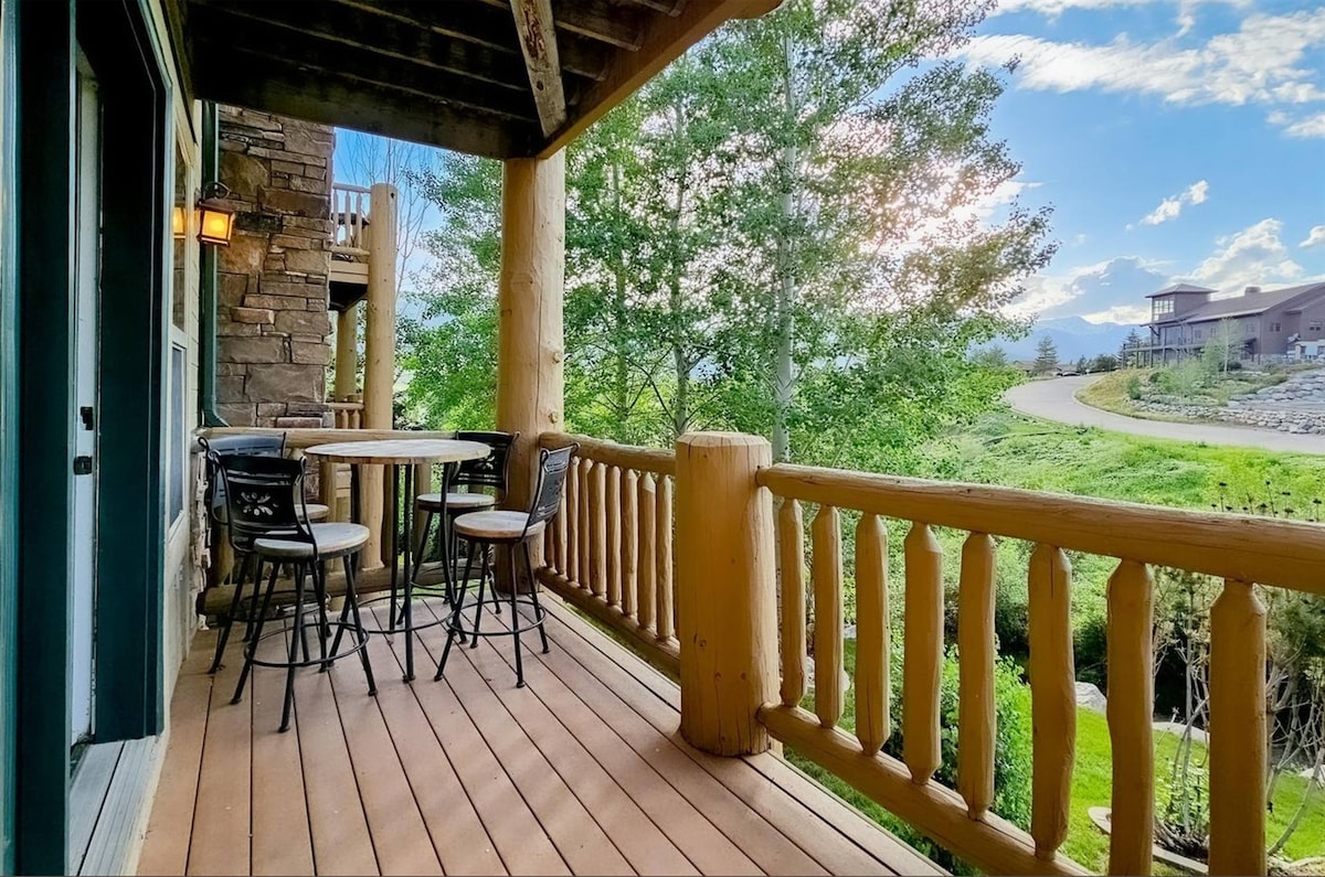 A wooden deck is shown, featuring a round table with two high-top chairs. The scene is framed by log railings and is surrounded by greenery, with a glimpse of a winding road in the background and distant mountains under a sunny sky.