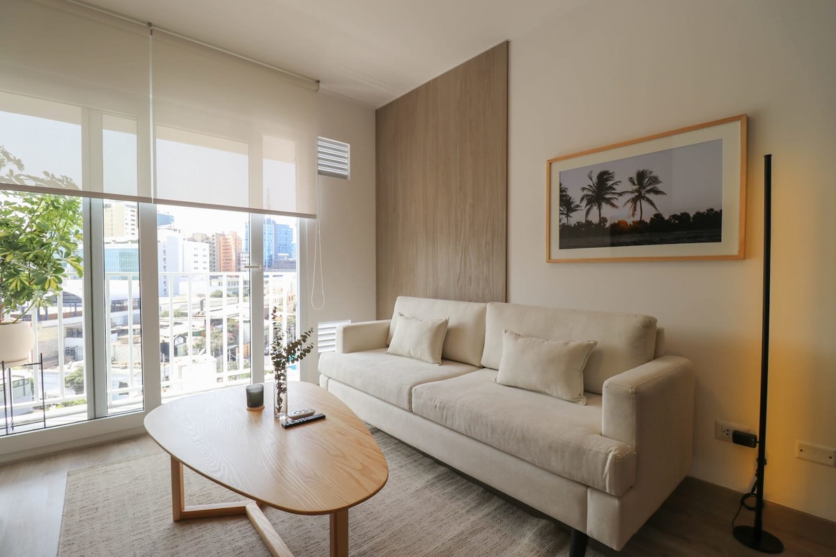 A modern living area is featured, with a light-colored sofa positioned across from a circular wooden coffee table. Large glass doors allow natural light to fill the space and provide access to a balcony with city views. A potted plant adds a touch of greenery.