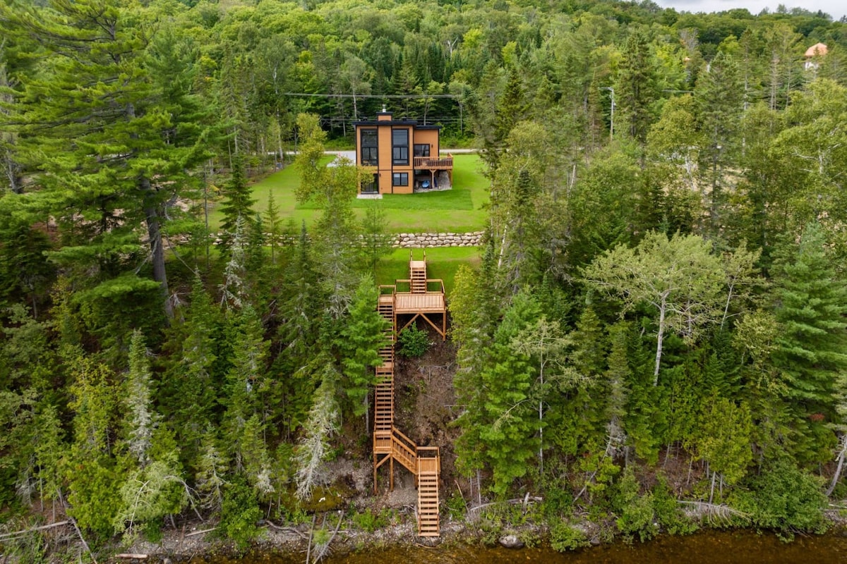 An aerial view reveals the chalet nestled among lush greenery, showcasing its modern architecture against the natural backdrop. The surrounding trees provide a sense of seclusion, while wooden stairs lead down to the water's edge, enhancing the connection to the serene lake.