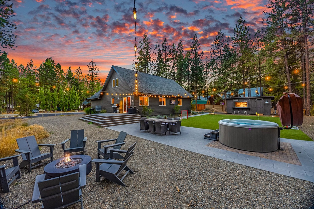 An outdoor space is highlighted by an illuminated fire pit surrounded by Adirondack chairs. A large hot tub sits nearby on a stone patio, with trees and a vibrant sunset in the background. The rustic home is visible, framed by twinkling string lights.
