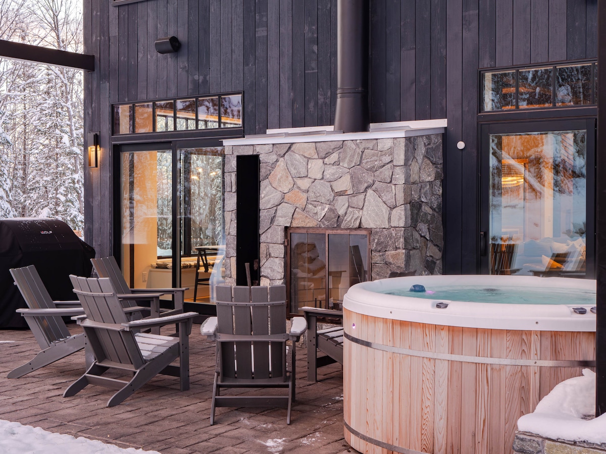 The outdoor area is highlighted by a classic Nordic hot tub beside a stone fireplace. Wooden lounge chairs are arranged for relaxation, while large windows offer views into the interior. The surrounding winter landscape is partially visible, creating a serene atmosphere.