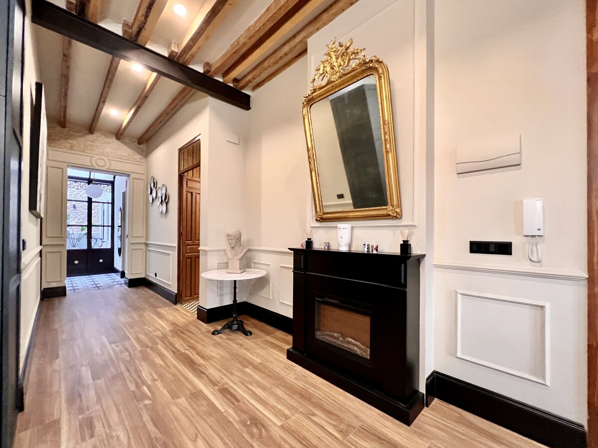 A spacious hallway features polished wooden flooring and high ceilings, highlighted by exposed wooden beams. A decorative mirror is elegantly framed and situated above a sleek electric fireplace. The entrance door is visible at the far end, surrounded by intricate wall details.