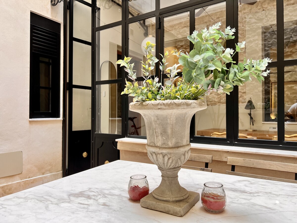 A stone vase filled with greenery sits atop a marble dining table. Soft candlelight from small glass holders adds warmth to the scene. Large glass doors provide a view of the indoor space, revealing a cozy atmosphere with natural light filtering through.