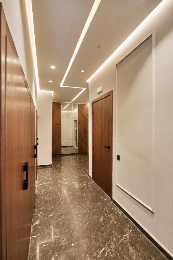 A well-lit hallway features glossy marble flooring and contemporary lighting along the ceiling. Wooden doors on either side lead to different rooms, while a large mirror reflects the space, enhancing the hallway's sense of depth and brightness.