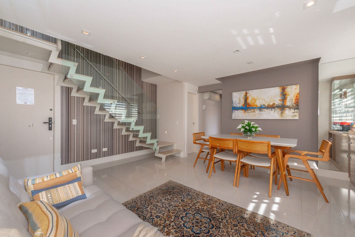 The spacious living area features a cozy sofa and a stylish dining table surrounded by modern chairs. Light-colored walls and smooth flooring enhance the welcoming ambiance, while an elegant staircase leads to the upper level. A decorative painting adds a touch of color to the room.