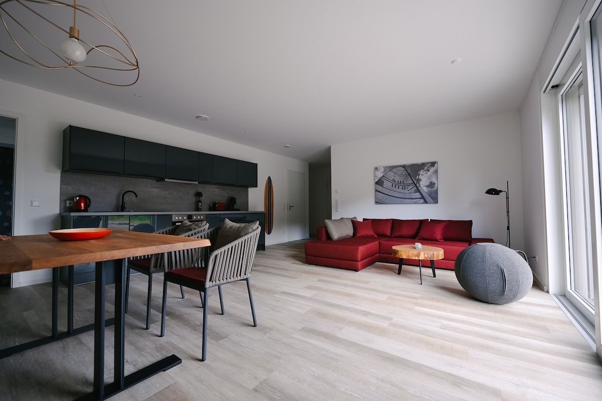 An open living area is shown, featuring a red sectional sofa and a round coffee table. A dining table with four chairs is positioned nearby. The kitchen area has modern cabinetry and appliances. Large glass doors allow natural light to brighten the space.