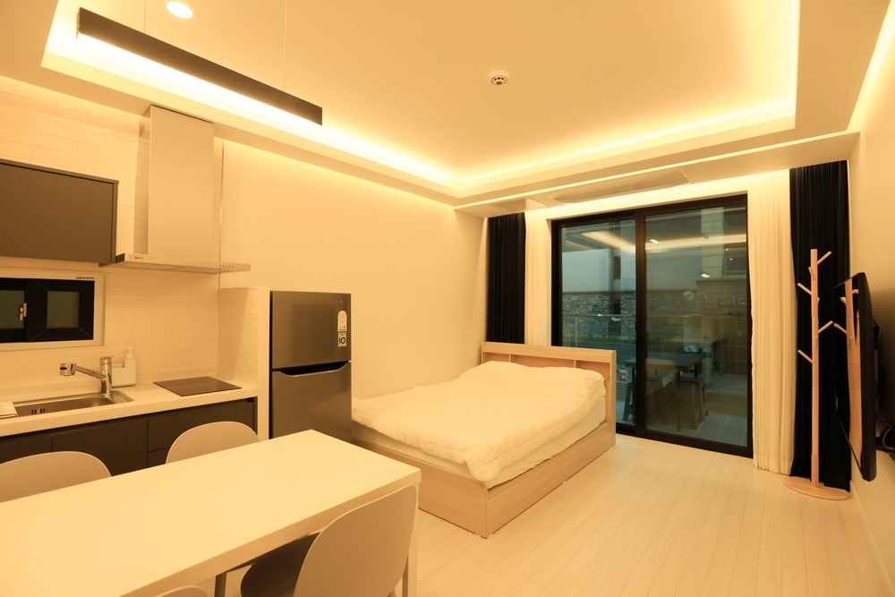 The studio room features a spacious bed with a light-colored comforter, complemented by soft lighting from the ceiling. A modern kitchenette and a table for four are visible, with large glass doors offering access to an outdoor area.