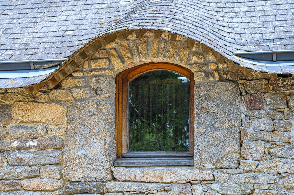 A rounded stone window is set into the rustic facade of the house, showcasing the texture of the aged stonework. The window reflects the greenery outside, creating a connection with the surrounding natural environment.