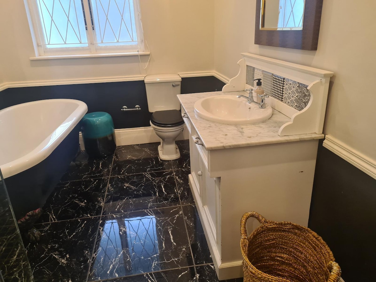 A well-appointed bathroom features a freestanding bathtub beside a modern toilet. A marble countertop is accompanied by a sink and a large mirror. The dark tiled floor complements the light walls, while a woven basket adds texture to the space.