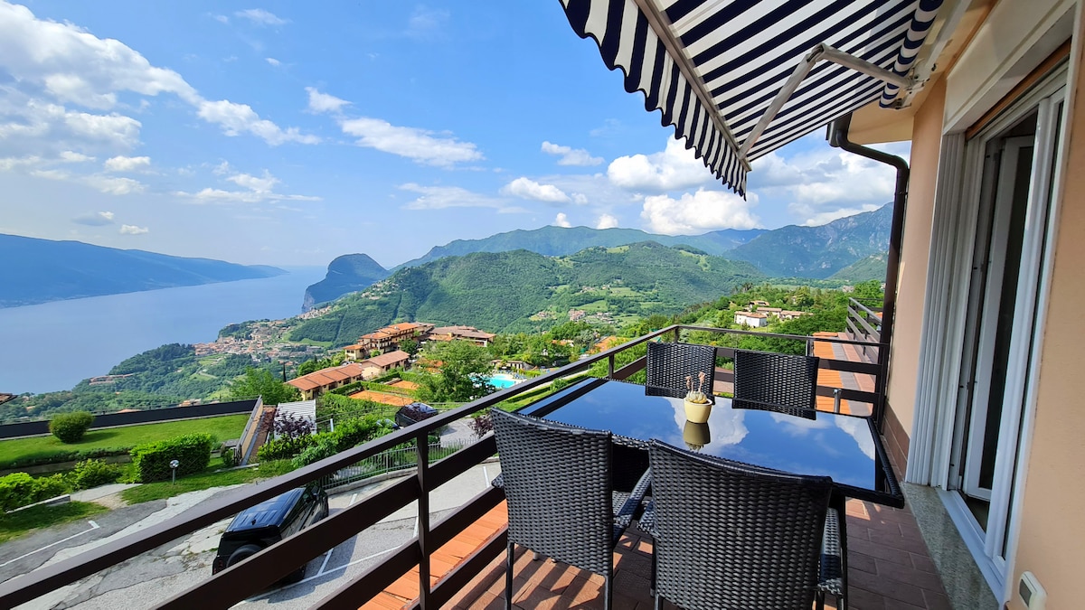 A furnished balcony overlooks a stunning lake view, framed by rolling hills and lush greenery. A dark wicker dining set with a glass top is positioned on the balcony, inviting outdoor relaxation. The sky is bright, and striped awnings provide shade.