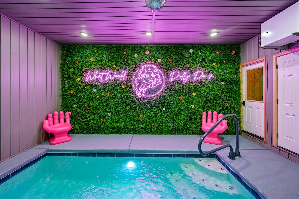 A vibrant indoor heated pool area features a striking green wall adorned with colorful flowers and neon pink signage. Two oversized pink chairs are positioned beside the pool, which is framed by a textured surface and a safety railing.