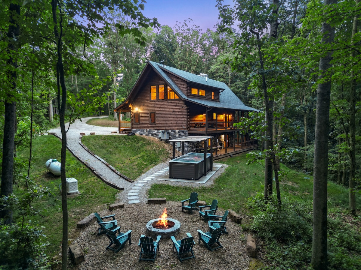 A spacious cabin nestled among trees features a stone fire pit surrounded by comfortable seating. A winding pathway leads to a covered swim spa situated beside the cabin, offering an inviting outdoor space for relaxation.