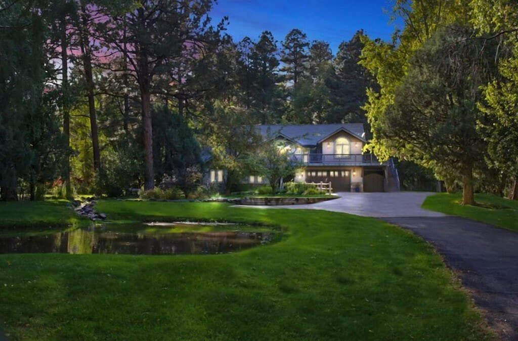 The expansive exterior of the Riverbend Hideaway is highlighted by lush greenery and a tranquil pond, set against the backdrop of towering trees. The illuminated house features a welcoming design, offering a glimpse of the garage and a spacious driveway winding through the manicured lawn.