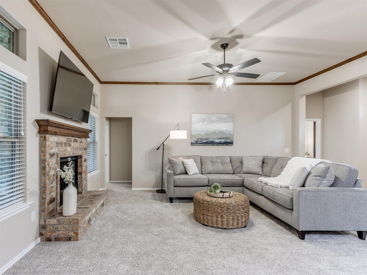 A spacious living room is featured with a large sectional sofa and a round coffee table. Natural light enters through multiple windows, and a mounted flat-screen TV is positioned above a brick fireplace. Soft carpeting and neutral-colored walls enhance the cozy atmosphere.
