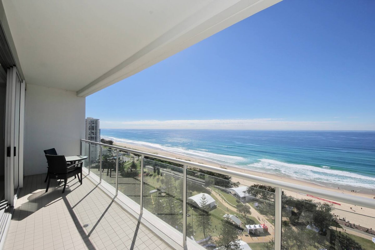 A private balcony extends from the apartment, featuring a chair and a table. Expansive views of the coastline and waves are visible, showcasing sandy beaches and gentle ocean currents under a clear blue sky.