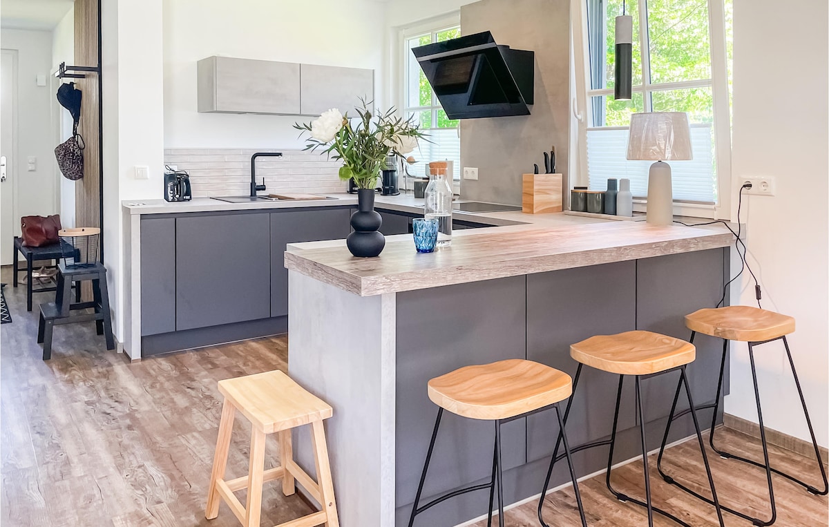 A modern kitchen features sleek cabinetry and an island with three wooden stools. Natural light fills the space through large windows, illuminating the minimalist design which includes a black extractor hood and stylish countertop decor.