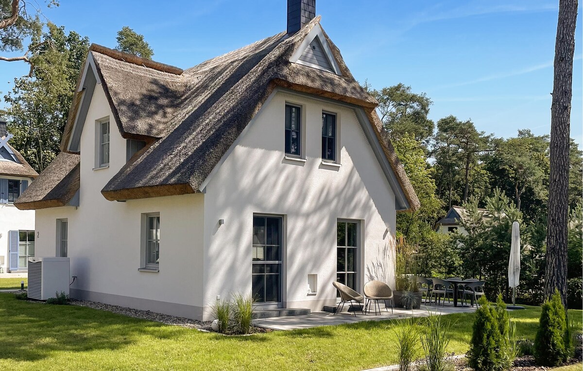 A charming holiday home with a thatched roof is surrounded by lush greenery. Sunlight casts gentle shadows on the white exterior, while a small seating area with modern chairs sits on the terrace, inviting relaxation in the outdoor space.