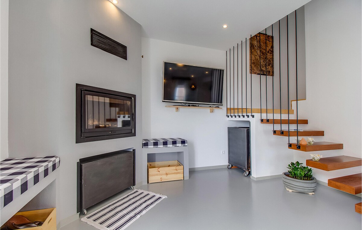 A modern living area is shown with a wall-mounted television above a sleek fireplace. Two striped benches provide seating, while a staircase with wooden steps leads to the upper floors. Natural light fills the space, contributing to the open feel.