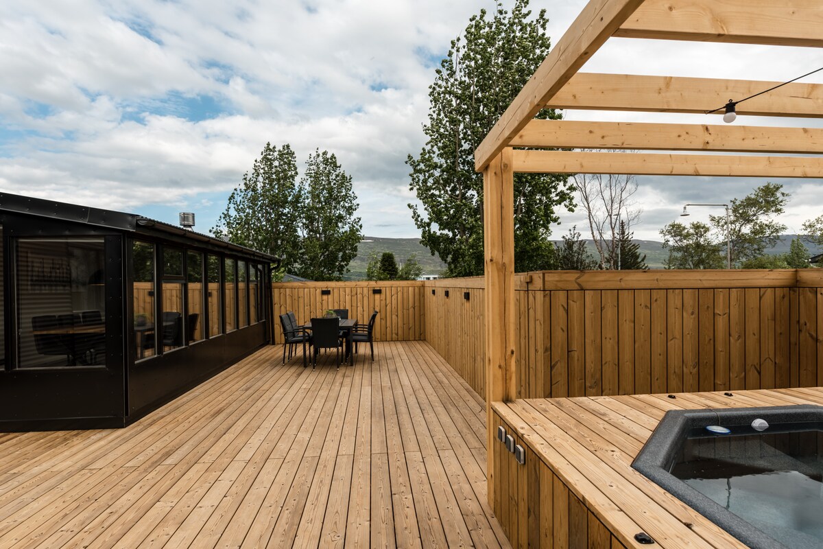 An expansive wooden deck is surrounded by tall fencing for privacy. A hot tub is integrated into the space, with outdoor furniture including a dining table and chairs positioned for relaxation. Green trees are visible beyond the deck, contributing to the serene environment.