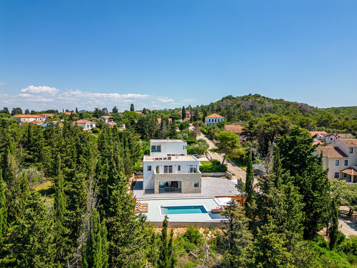 An aerial view of Villa Molaris surrounded by lush greenery and tall trees. The villa features a spacious outdoor area with a swimming pool. Nearby homes and winding paths create a tranquil village setting against a clear blue sky.