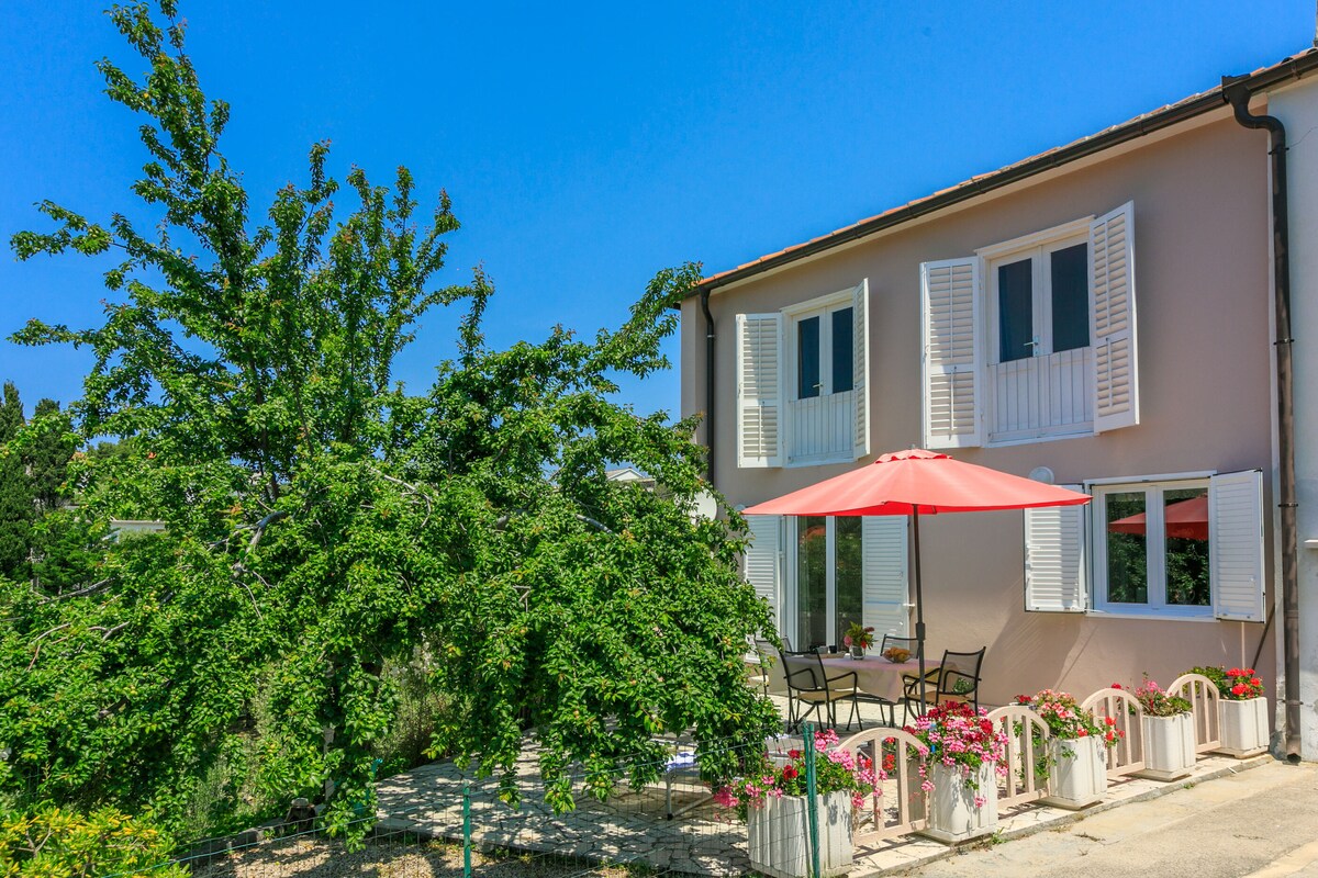 A comfortable outdoor seating area is situated beneath a large umbrella, featuring a table and chairs surrounded by potted flowers. The property features a two-story structure with multiple windows adorned with shutters, complemented by a lush tree nearby and clear blue skies.