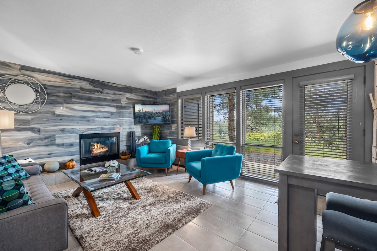The living area features a modern fireplace surrounded by a wood panel accent wall. Two teal chairs provide comfortable seating, complemented by a neutral rug under a coffee table. Large windows allow natural light to fill the space, with views of the scenic outdoors.