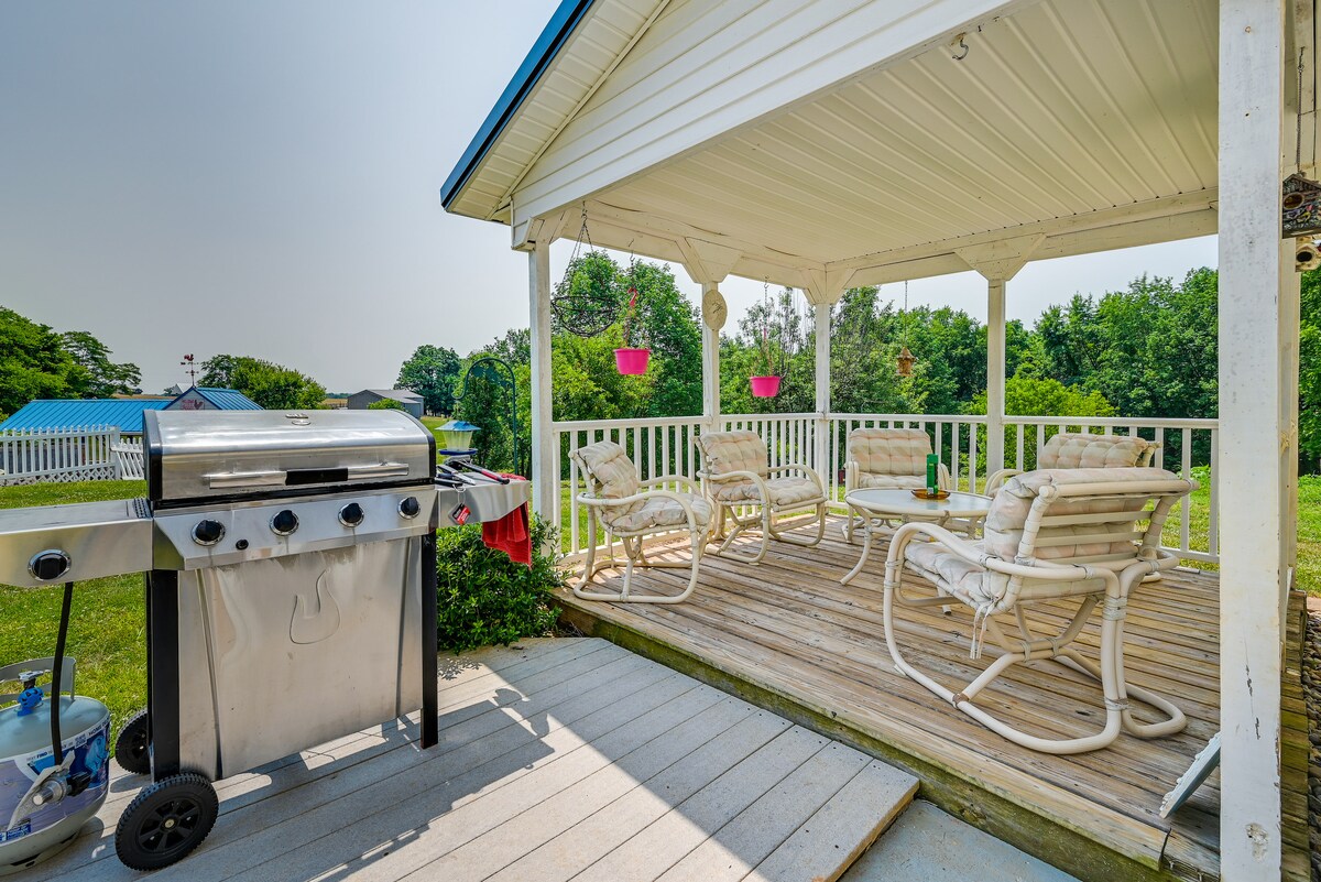 A covered outdoor deck area features a gas grill and multiple white armchairs surrounding a wooden table. Greenery is visible in the background, complemented by hanging planters, creating a relaxed outdoor space.