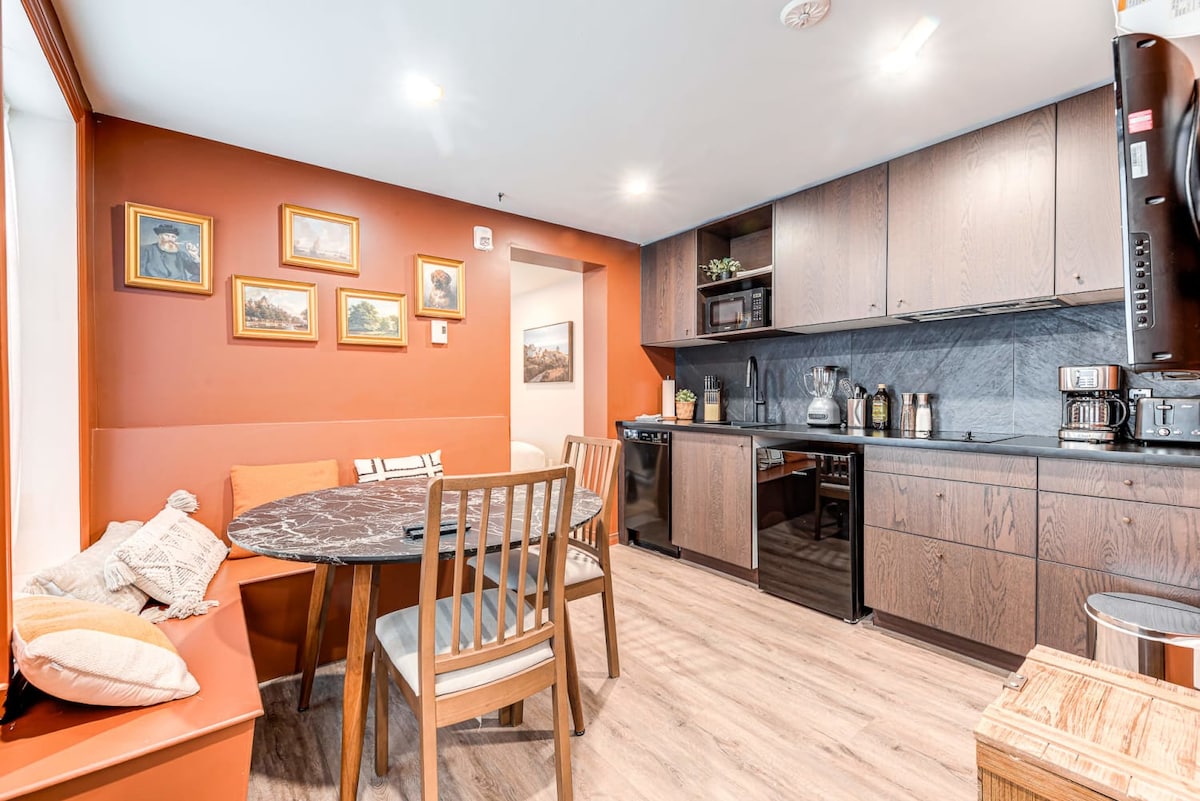 A compact kitchen area is visible, featuring modern cabinetry and a dark countertop. A round dining table with four wooden chairs is positioned against a warm, orange wall adorned with framed artwork. Light streams in through a nearby window, illuminating the space.
