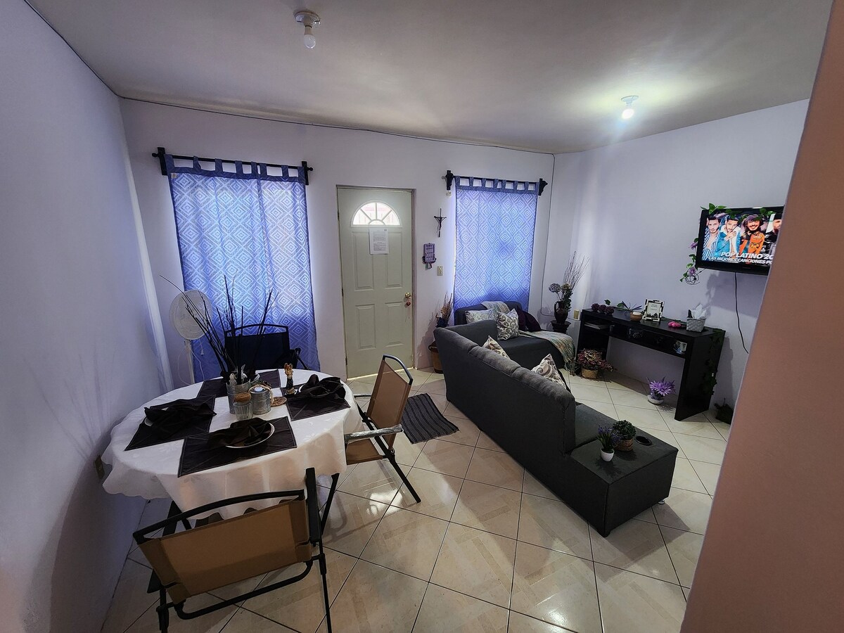A living area is furnished with a dark sofa and a small round dining table with metal chairs. Light filters through sheer curtains at the windows. A television is positioned on a stand against the wall, adding to the comfortable ambiance of the space.