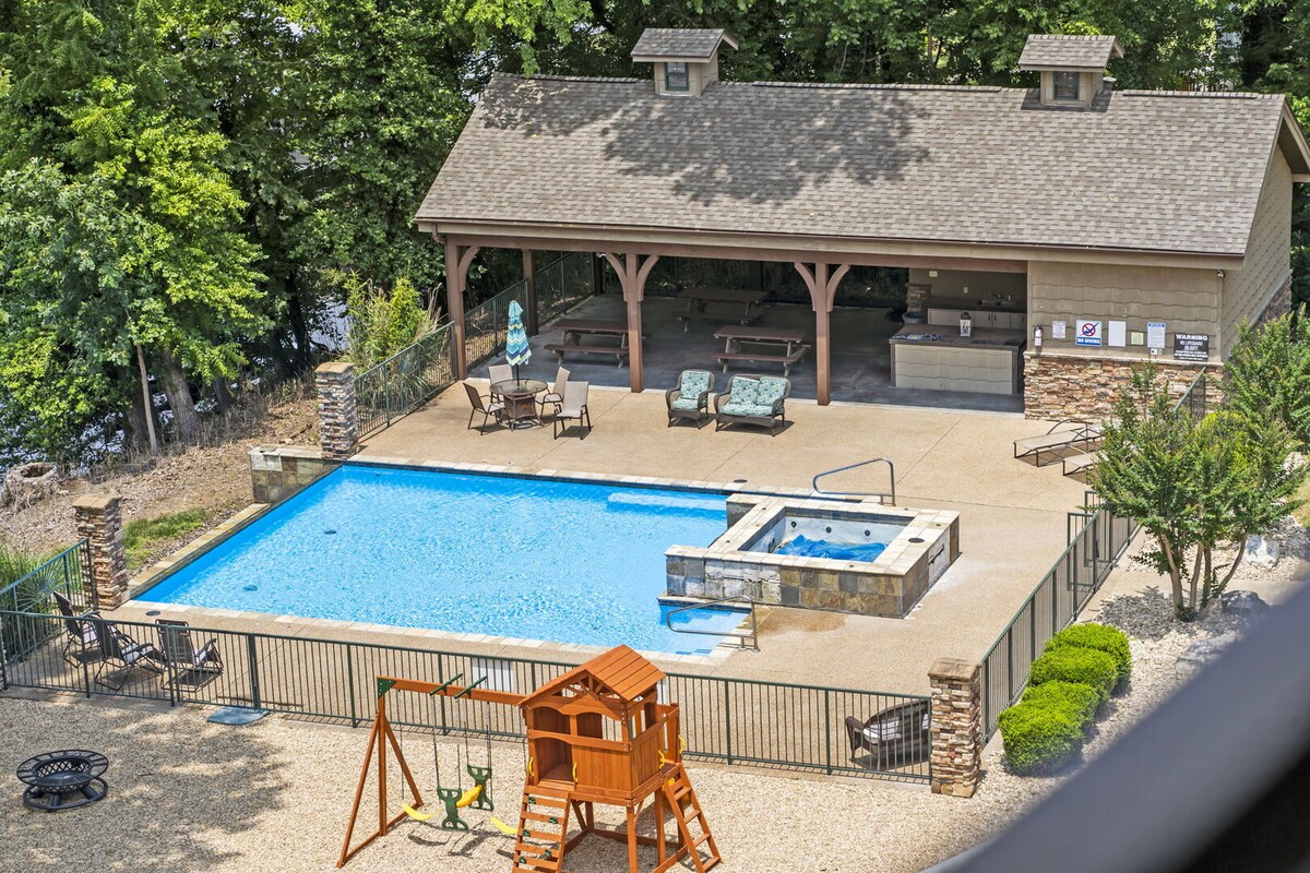 An outdoor pool area is surrounded by a security fence, featuring a spacious swimming pool and a hot tub. A shaded pavilion offers seating and amenities, while nearby, a playground set invites families to enjoy outdoor activities.