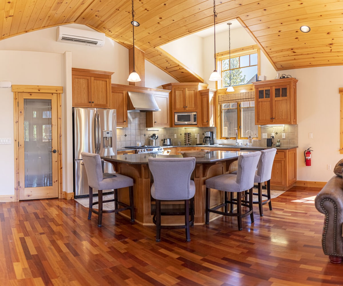 A warm kitchen area features granite countertops and a central island with bar-style seating for four. Natural light floods the space through large windows, highlighting the wooden cabinets and floors. Stainless steel appliances are seamlessly integrated into the design, creating a functional cooking environment.