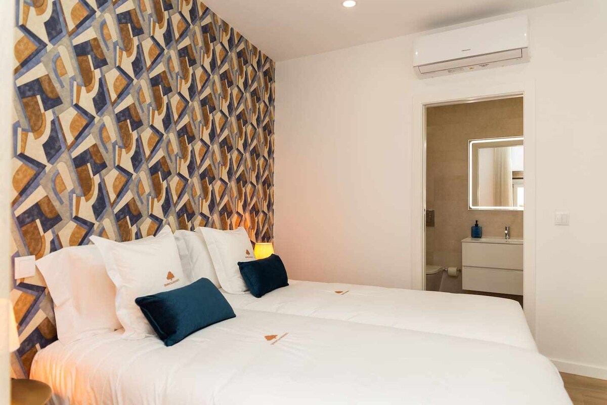 A bright sleeping area features two spacious beds with crisp white linens. Decorative pillows with deep blue accents provide contrast against the textured wallpaper. A doorway leads to a bathroom area, where modern fixtures can be seen in the reflection of the mirror.
