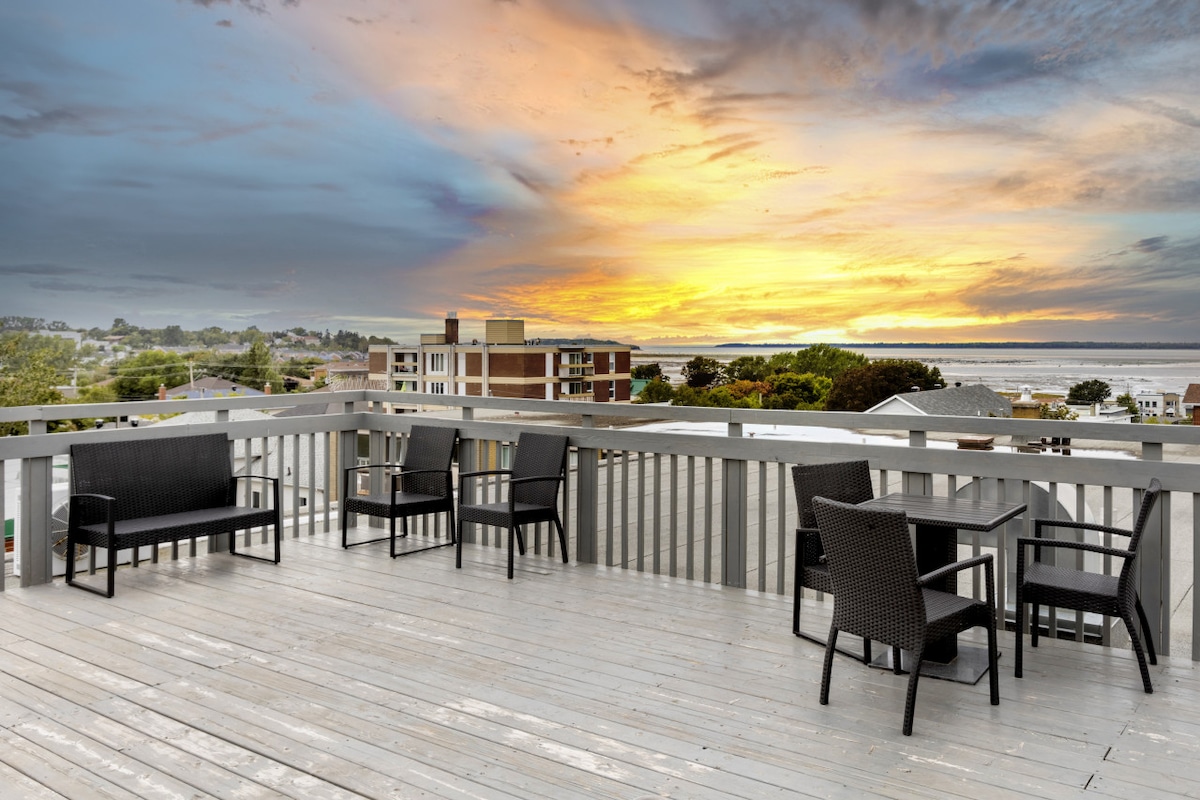 A spacious rooftop terrace features a seating arrangement with black wicker furniture, including a table and several chairs. The sunset creates a colorful sky above, while the view includes distant buildings and the coastline.