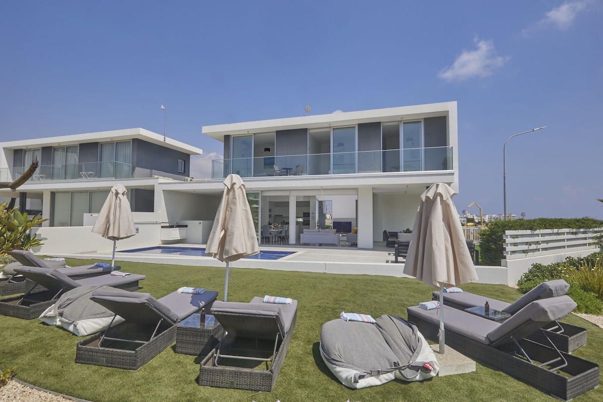 A contemporary villa is shown with a spacious terrace and a landscaped garden. Sun loungers are arranged around a private pool, complemented by large umbrellas for shade. Expansive glass doors provide a view into the open living space. The overall layout promotes relaxation and outdoor enjoyment.